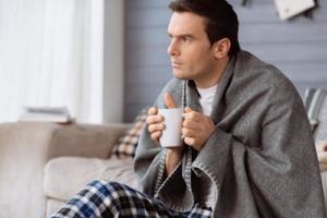 a person sits on a couch in a blanket sipping tea possibly dealing with alcohol withdrawal symptoms