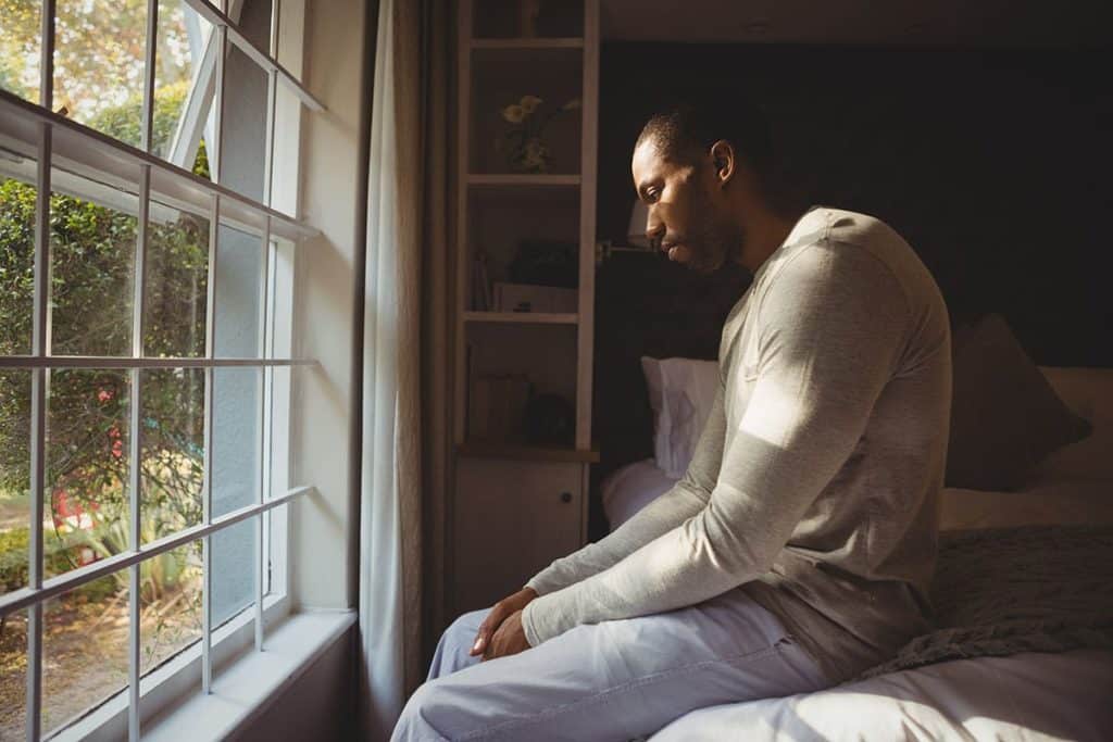 a person experiencing meth addiction symptoms sits sadly on the edge of a bed looking out of a window